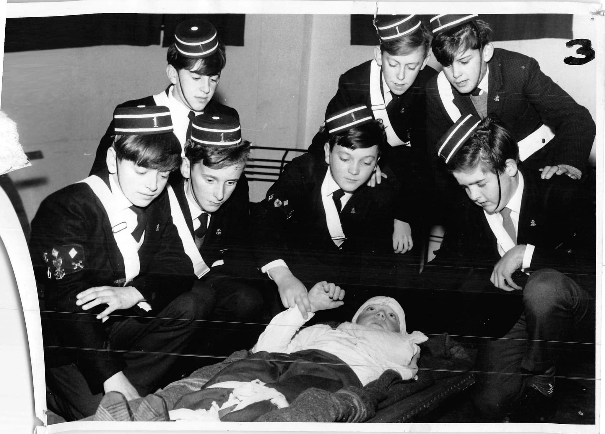 A black and white photo showing 7 Boys Brigade members in uniform surrounding a bandaged boy lying on the floor, the boy on the floor is raising his hand towards a concerned member.