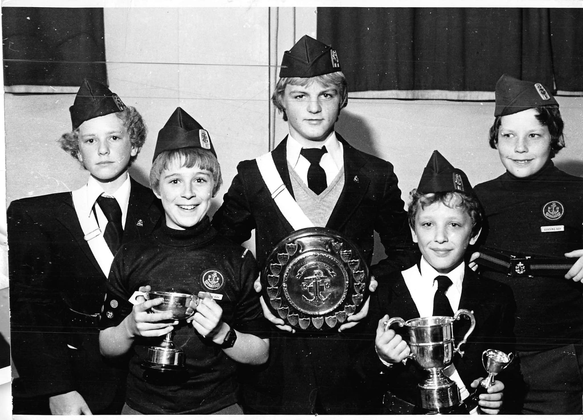 A black and white photo of 5 boys brigade members, the central member is holding a big disc trophy whilst the other 4 all have small trophies.